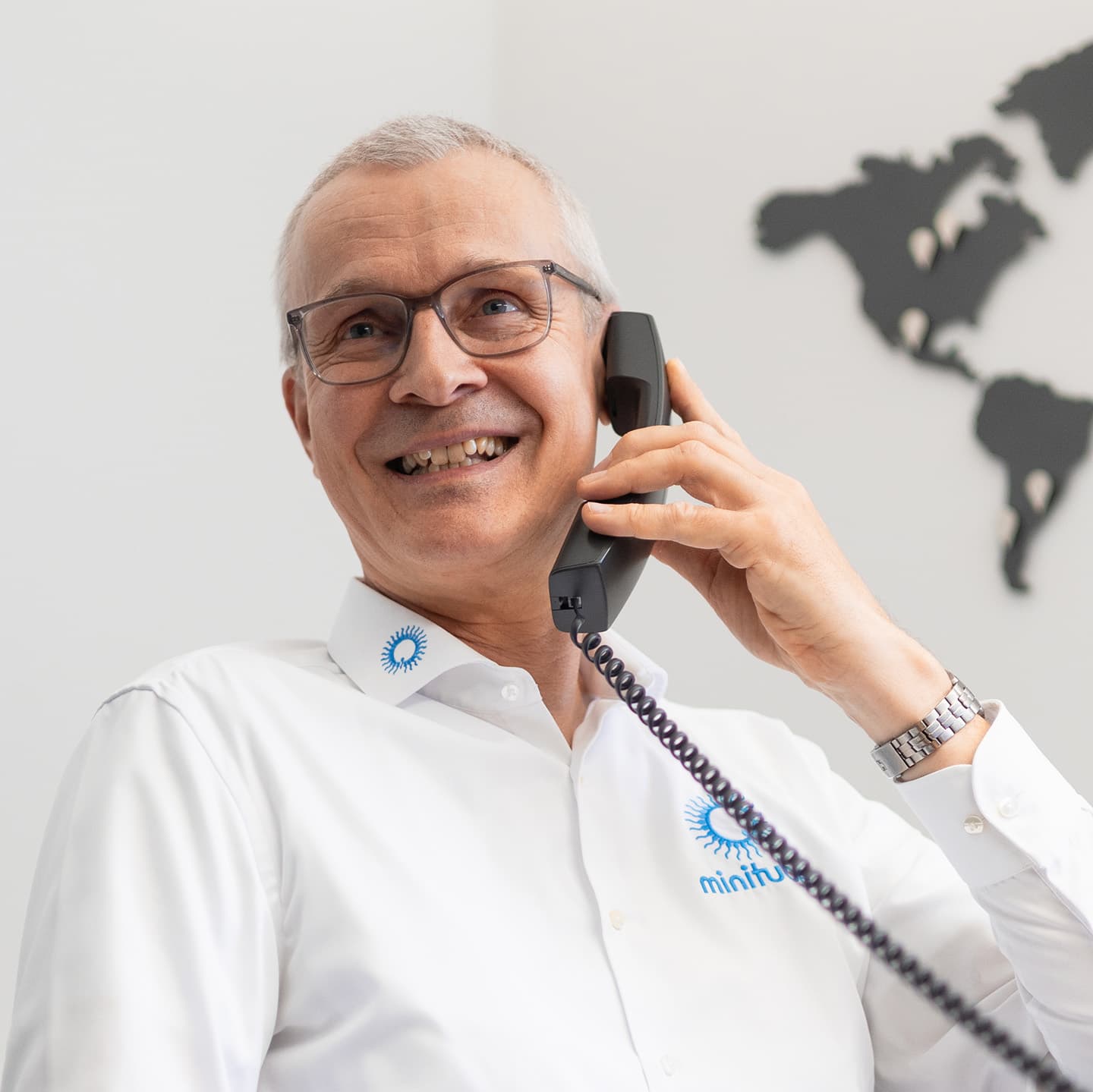 Professional lifestyle photo of Paul Gassner smiling while talking on a black office telephone receiver, wearing a white branded Minitube shirt.
